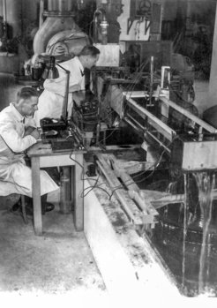Two people in white coats work at a table with technical measuring instruments as water flows into a basin.